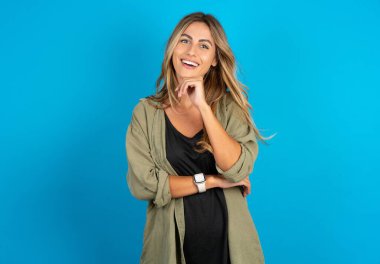 beautiful blonde woman wearing overshirt on blue background laughs happily keeps hand on chin expresses positive emotions smiles broadly has carefree expression clipart