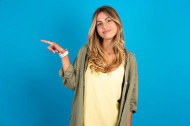 beautiful blonde woman wearing overshirt on blue background points to side on blank space demonstrates advertisement. People and promotion concept clipart
