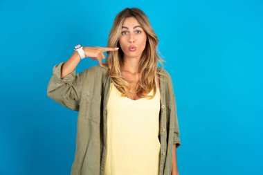 Güzel, sarışın bir kadın, mavi arka planda tişört giyiyor, dudaklarını işaret ediyor, işaret parmağıyla işaret ediyor, ameliyattan sonra etkisini gösteriyor.,