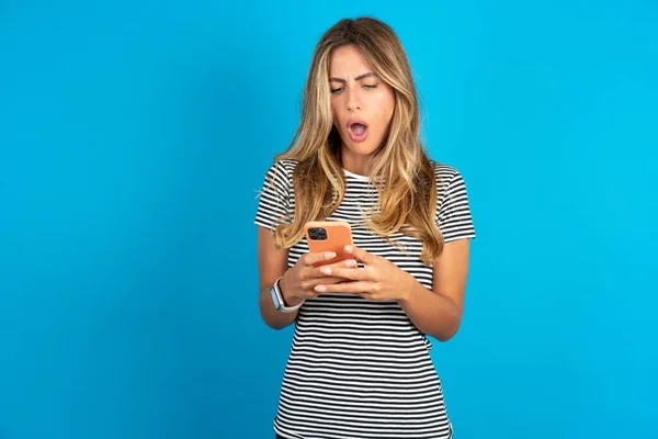 スマートフォンの読書ソーシャルメディアニュースや重要な電子メールを使用して青い背景にストライプTシャツを着ている若い美しい女性を驚かせます — ストック写真