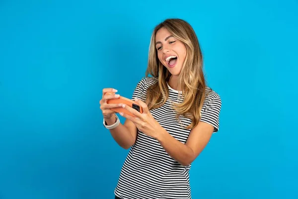 携帯電話でゲームをする青い背景の上にストライプTシャツを着ている興奮する若い美しい女性の肖像画 — ストック写真
