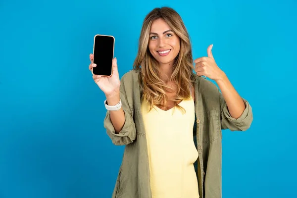 漂亮的金发女人穿着蓝色背景的衬衫显示空白屏幕智能手机 推荐新的应用程序 — 图库照片