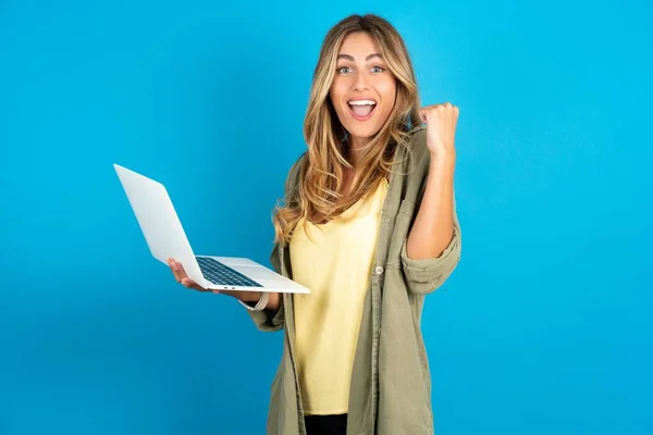 stock image beautiful blonde woman wearing overshirt on blue background hold computer open mouth rise fist