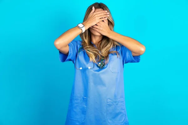 青いスタジオの背景の上に立っている美しい医師の女性は 手で目と口を覆い 驚きとショックを受けました 感情を隠す — ストック写真