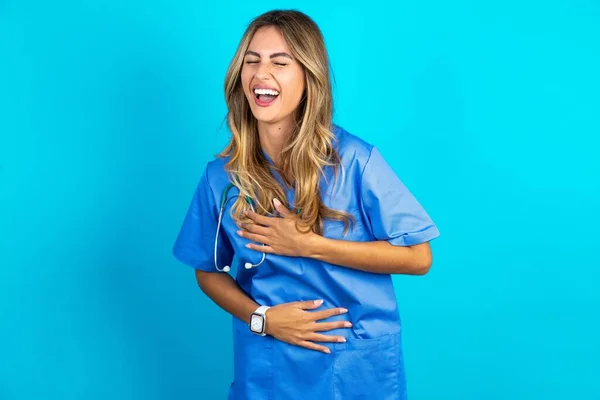 美しい医師の女性が青いスタジオの背景に立って笑って ボディ上の手で面白いクレイジーな冗談のために大声で笑います — ストック写真