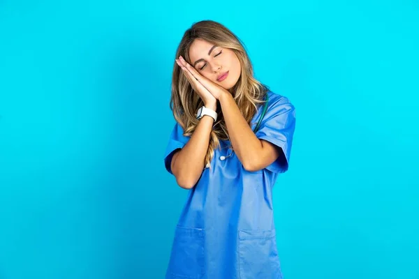 青いスタジオの背景の上に立っている美しい医師の女性 疲れた夢を寝て 目を閉じて微笑んでいる間一緒に手でポーズ — ストック写真