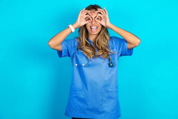青いスタジオの背景の上に立っている美しい医師の女性は 舌を突き出し 指を通して見ているような良いジェスチャーをする クレイジー表現 — ストック写真