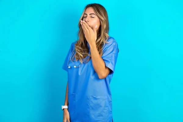 青いスタジオの背景の上に立っている美しい医師の女性は 仕事で一日中過ごした後 疲れています — ストック写真