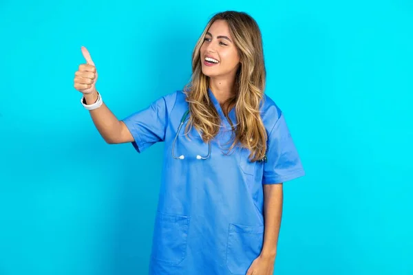 美丽的医生女士站在蓝色工作室的背景上看起来很自豪 微笑着向旁边做着竖起大拇指的手势 干得好 — 图库照片