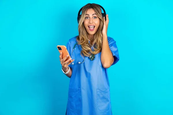 站在蓝色工作室背景上的积极漂亮的医生女人拿着连接着耳机的现代手机 从美好的情感中握紧拳头 欢呼雀跃 — 图库照片