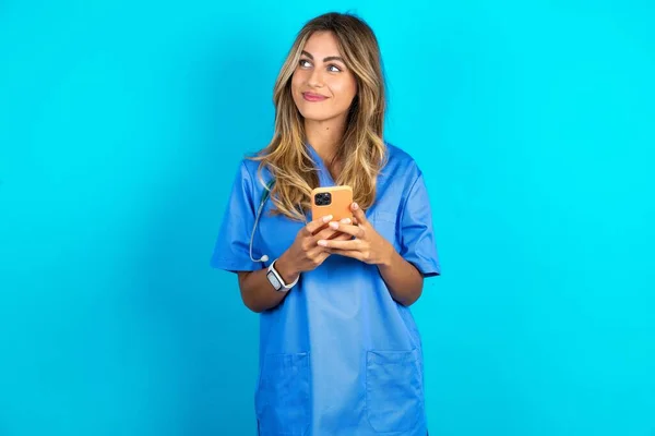 美丽的医生女士站在蓝色工作室的背景上 手持手机 带着梦幻般的神情望着旁边 — 图库照片