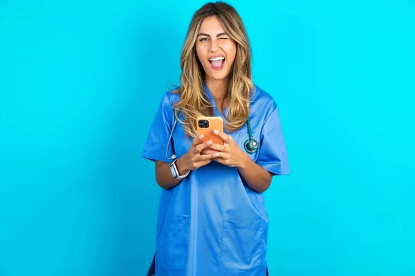 兴奋美丽的医生女人站在蓝色工作室的背景下眨眼 用智能手机看社交网络新闻 — 图库照片