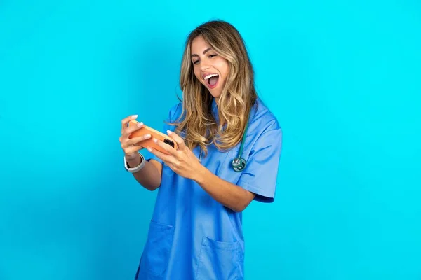 一个兴奋美丽的医生女人站在蓝色工作室背景下用手机玩游戏的画像 — 图库照片