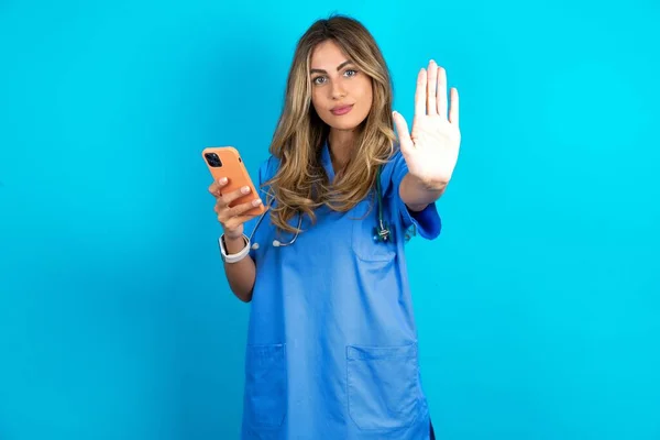 美丽的医生女士站在蓝色工作室的背景上 用智能手机发短信 张开双手 做着严肃而自信的手势 做着防御手势 — 图库照片