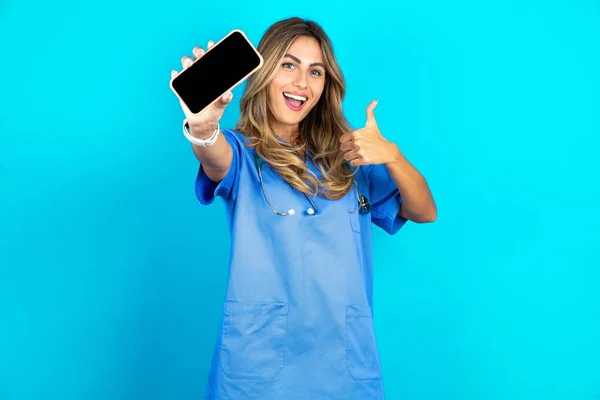 照片中美丽的医生女士站在蓝色工作室的背景上 手持手机 将黑屏大拇指伸出来 — 图库照片