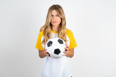 Genç ve güzel bir kadın futbol topunu beyaz arka planda tutuyor. Parmaklarıyla işaret ediyor. Reklam yapıyor, şaşırıyor ve ağzını açıyor.