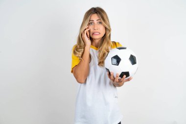 Korkmuş ve güzel bir kadın futbol tişörtü giymiş beyaz arka planda telefon ve futbol topu tutuyor.
