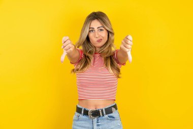 Sarı stüdyo arka planında çizgili tişört giyen güzel, sarışın bir kadın iki eliyle baş parmağını aşağı gösterdiği için üzgün. Konseptten nefret.