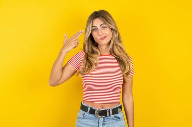 Sarı arka planda çizgili tişört giyen mutsuz sarışın kadın intihar jesti yapıyor ve tabancayı eliyle taklit ediyor, dudaklarını kıvırıyor, iki parmağını şakakta tutuyor.