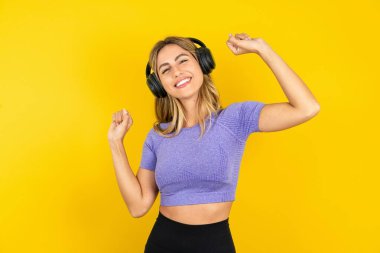 Stüdyonun sarı arka planında spor kıyafetleri giyen kaygısız genç bir kadın dişlek bir gülümsemeyle kol dansı yapıyor müziğin ritmiyle tasasız hareketler yapıyor, müzik listesinden kulaklıkla müzik dinliyor.