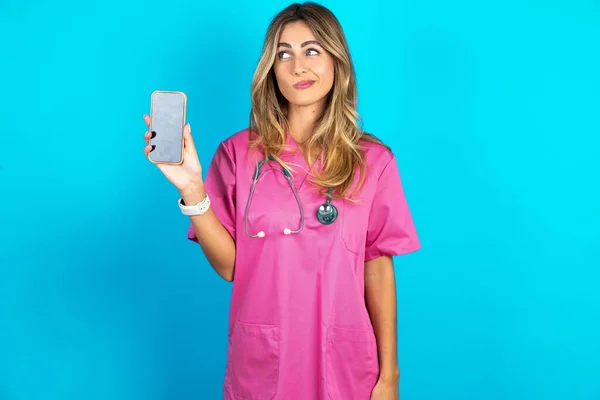 带听诊器身穿粉色军服的高加索女医生手持新手机 看上去神秘极了 旁边是现代手机的空白显示屏 — 图库照片