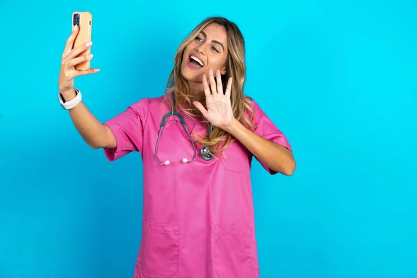 带听诊器身穿粉色医疗制服的高加索女医生手持现代手机 用手势向人们问好 人民现代技术概念 — 图库照片