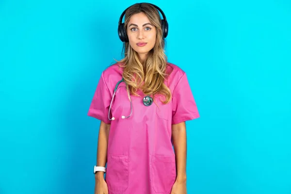 Serious displeased  caucasian woman doctor in pink medical uniform with stethoscope looks puzzled at camera being angry wears stereo headphones listens music