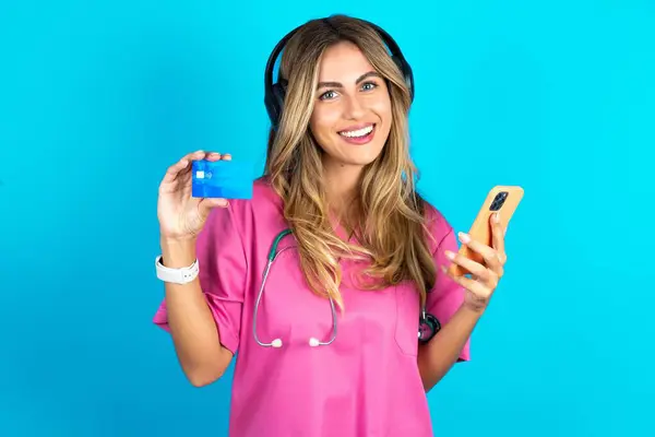 耳にステレオヘッドフォンを身に着けているピンクの医学の制服の白人女性の医者は現代携帯電話およびクレジット カードを握ります — ストック写真