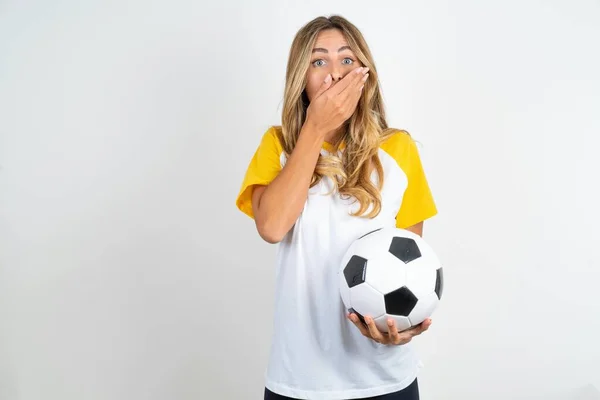白い背景の上にフットボールボールを保持する驚いた若い美しい女性は 両手を口に覆い 驚くべき何かを怖がります — ストック写真