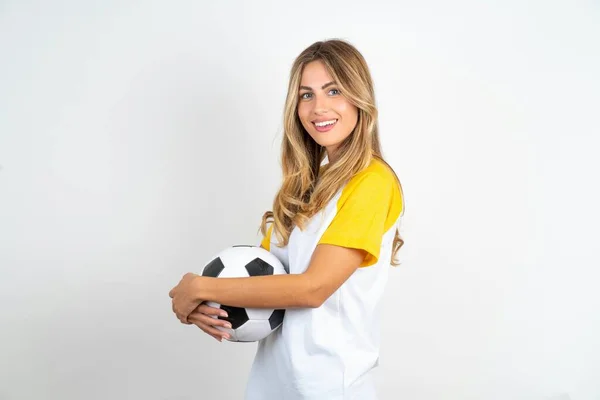 サッカーボールと笑顔を保持する折り畳まれた腕で立っている白い背景の上にフットボールTシャツを着ている美しい女性の肖像画 — ストック写真