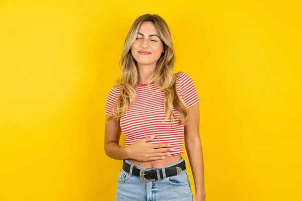 黄色いスタジオの背景にストライプTシャツを着ている美しい金髪の若い女性は 穏やかに微笑み 食べて満足のコンセプトに触れます — ストック写真