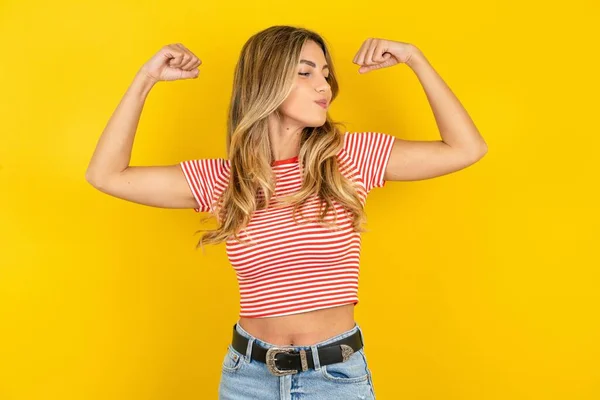 漂亮的金发女青年穿着条纹T恤 背景是黄色的工作室 露出胳膊肌肉 笑着自豪 健康概念 — 图库照片