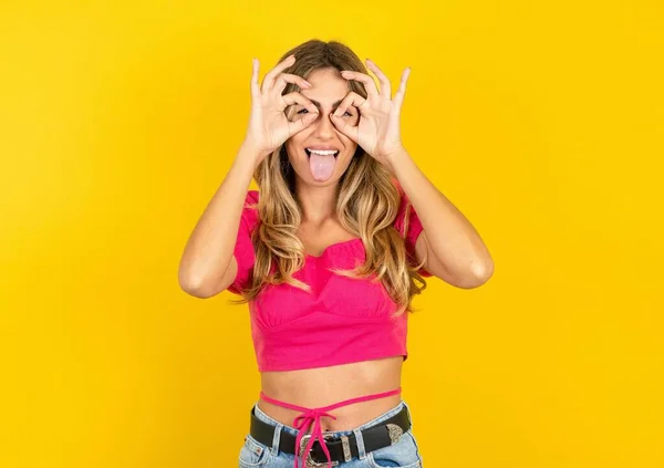 金发女人头戴粉色的庄稼顶盖 黄色背景 像双筒望远镜一样做手势 伸出舌头 眼睛透过手指看过去 疯狂的表情 — 图库照片