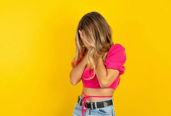 stock image Sad blonde young woman wearing pink crop top on yellow background crying covering her face with her hands.