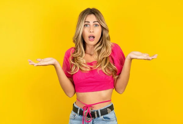 stock image Frustrated blonde young woman wearing pink crop top on yellow background feels puzzled and hesitant, shrugs shoulders in bewilderment, keeps mouth widely opened, doesn't know what to do.