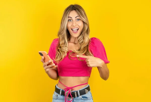 漂亮的金发碧眼的年轻女子手牵着新智能手机 头戴粉色麦片 背景是黄色的 — 图库照片