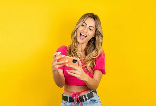 一个兴奋的金发碧眼的年轻女人穿着粉色的庄稼顶部黄色背景在手机上玩游戏的画像 — 图库照片
