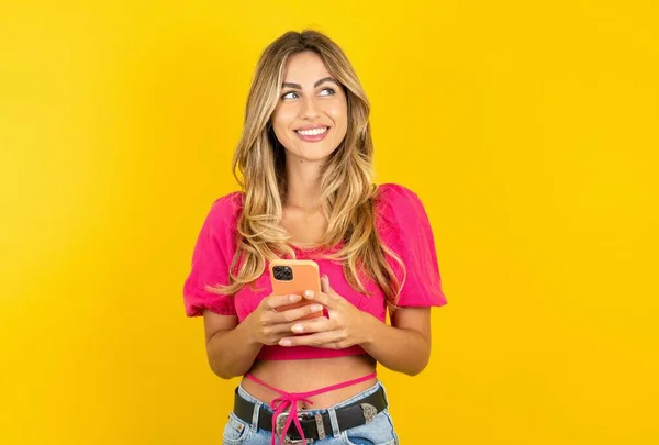 笑着金发碧眼的年轻的高加索女人穿着粉色的庄稼顶部黄色背景手牵着电话读好的青春新闻看空旷的空间广告 — 图库照片