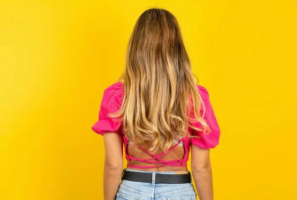 黄色の背景にピンクの作物の上を着用しているブロンドの若い女性のバックビュー スタジオショット — ストック写真