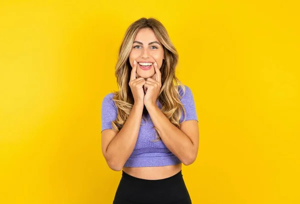 黄色いスタジオの背景の上にスポーツウェアを着ている幸せなブロンドの女性は 口の近くに指の指を保ち 陽気な笑顔を強制します — ストック写真