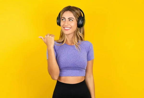 漂亮的金发女人穿着运动服站在黄色工作室的背景上 通过无线耳机听着音乐 然后把手指移开 在广告中复制空间 — 图库照片