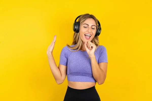快乐的金发女子穿着运动服在黄色工作室的背景上唱着最喜欢的歌 手牵着手 好像话筒戴着无线耳机 听着音乐 — 图库照片