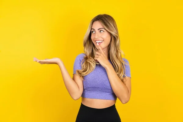 黄色いスタジオの背景にスポーツウェアを着用するポジティブな若い女性がプロモーションと指の歯に触れる — ストック写真