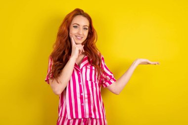 Sarı arka planda pembe pijama giyen komik kızıl saçlı kadın yeni bir ürünü açık havada tutuyor. Harika bir öneri.