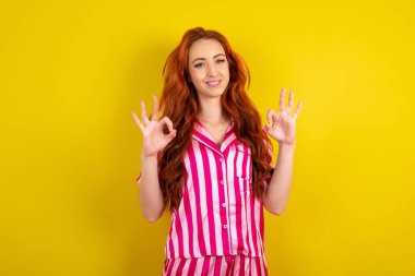 Sarı stüdyo arka planında pembe pijama giyen kızıl saçlı kadın iki elini de OK işaretinde gösteriyor. Onaylama veya öneri kavramı