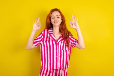 Sarı arka planda pembe pijama giyen kızıl saçlı kadın rahatla ve gözleri kapalı parmaklarıyla meditasyon hareketi yaparken gülümse. Yoga konsepti.