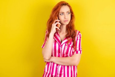 Sarı arka planda pembe pijama giyen üzgün kızıl saçlı kadın akıllı telefondan konuşuyor. İletişim konsepti.
