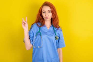 Genç, kızıl saçlı doktor kadın barış jesti yapıyor. Dudakları kapalı, V işaretini gösteriyor. Vücut dili kavramı