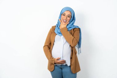 Müslüman hamile kadın tesettürlü kameraya kendinden emin bakıyor kollarını kavuşturmuş ve çenesini kaldırmış gülümsüyor. Olumlu düşünüyorum..