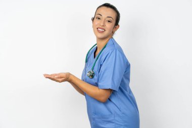 Arap doktor kadın mavi üniforma giymiş, elleri açık, fotokopi alanını gösteren, gülümseyerek mutlu bir gülümseme sunan.